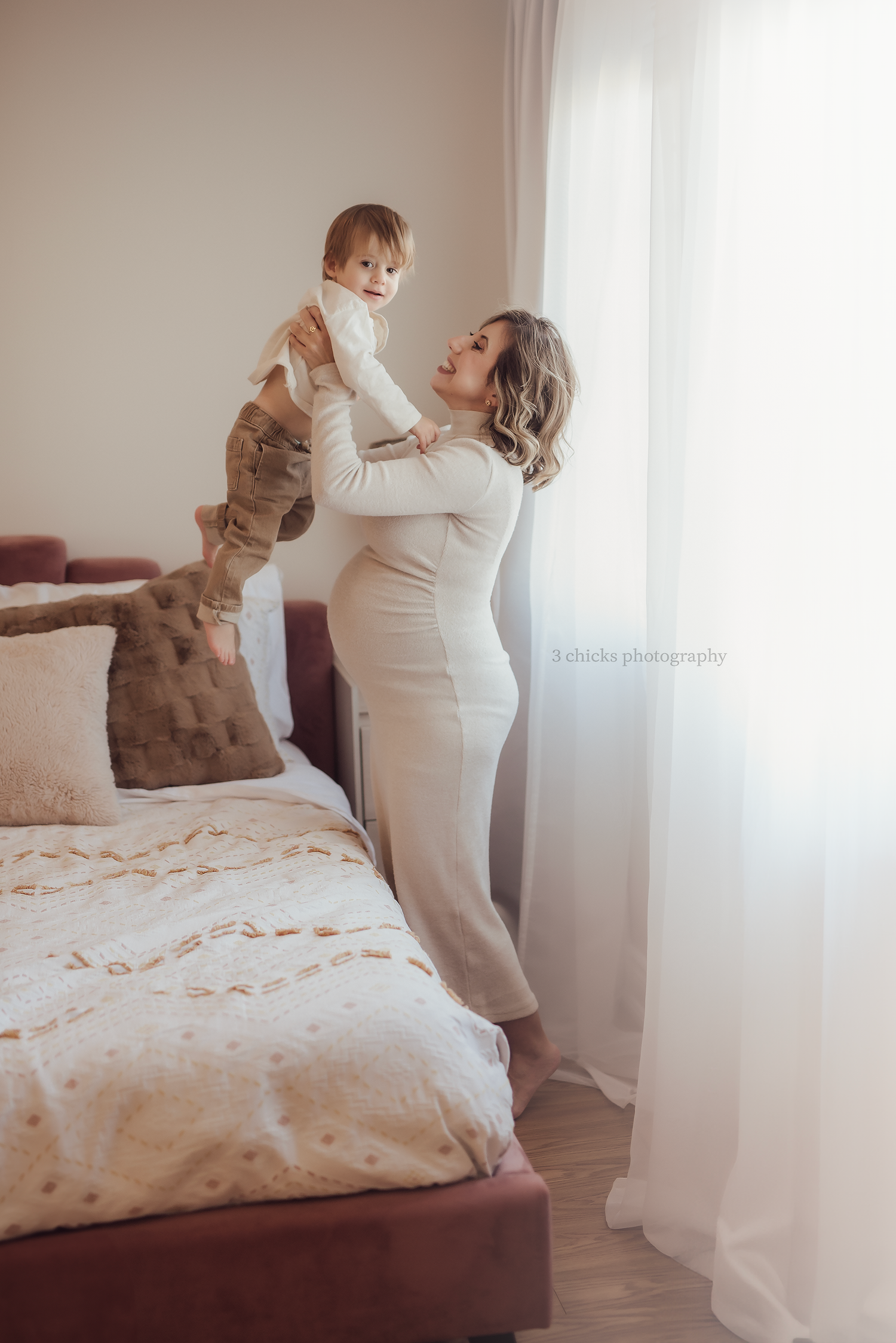 A mother in a white dress joyfully lifts her toddler in a bright, cozy bedroom during a maternity photo session.
