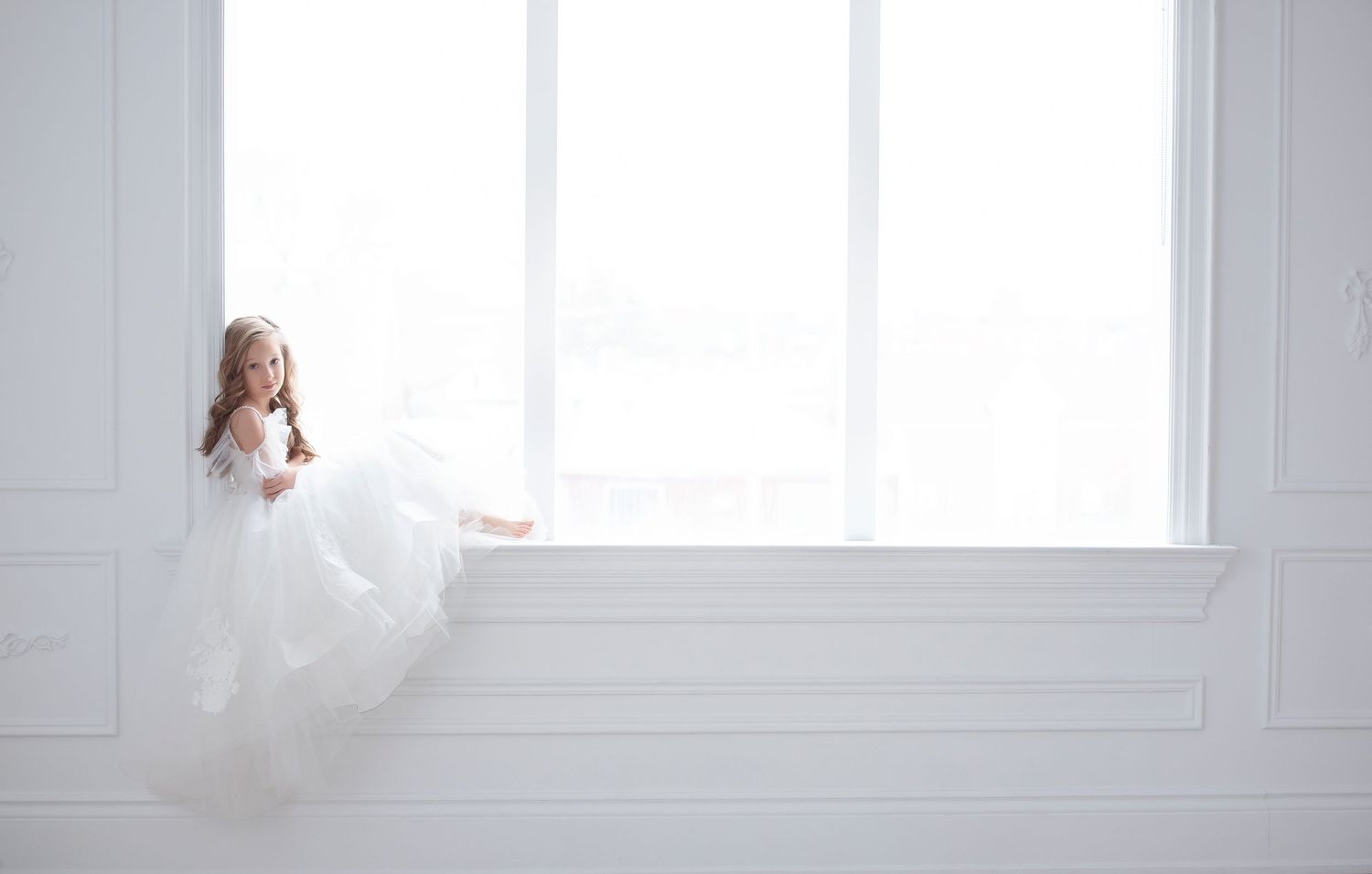 Young girl in white tulle dress sitting on window sill in bright white room.