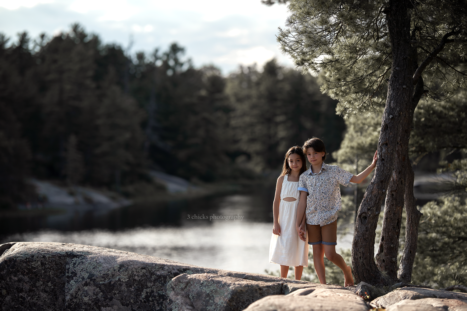 Two children in white outfits standing on rocks near a serene forest lake beside a pine tree.