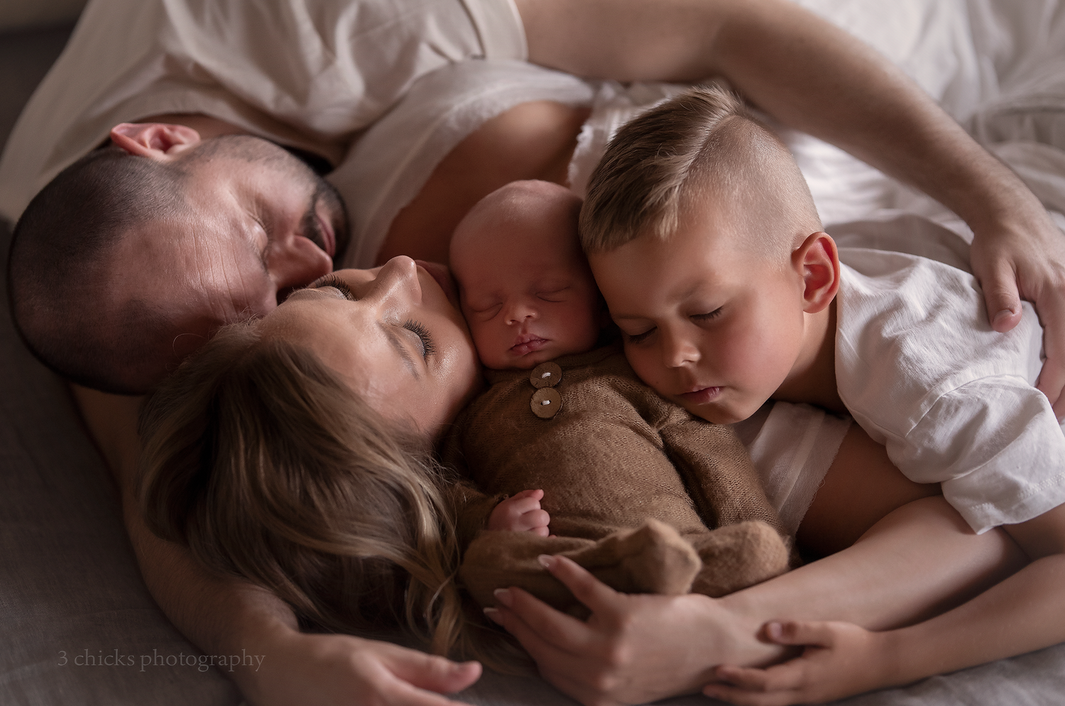 Family of four lying together on a bed, parents cuddling newborn baby and toddler in a tender moment.