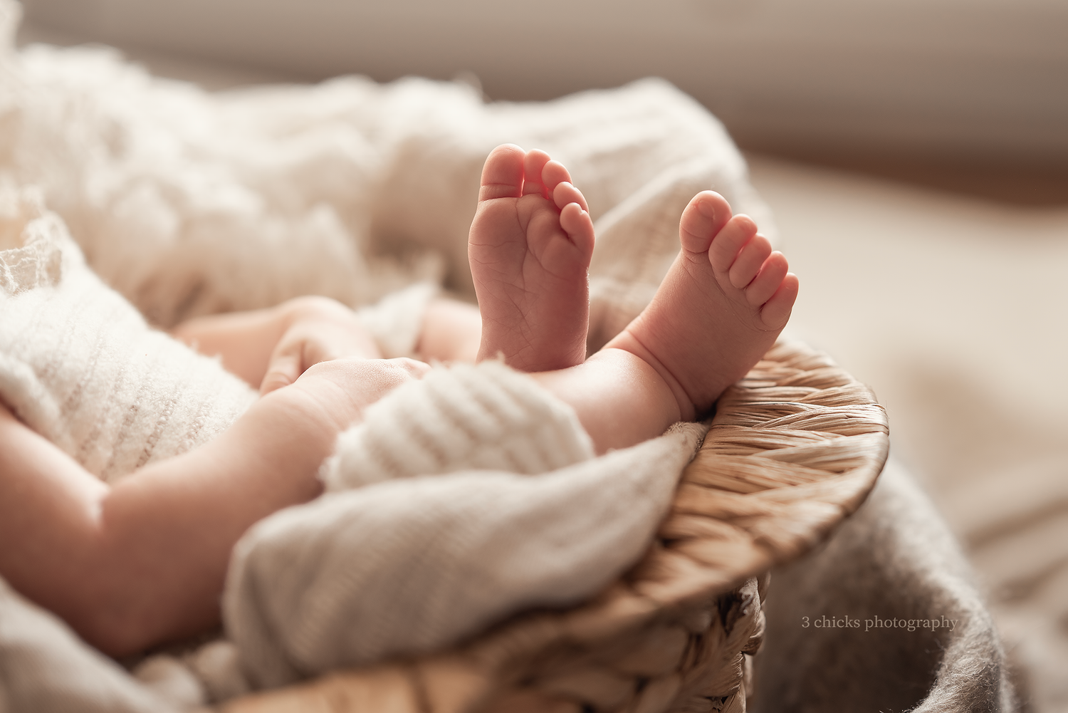 Tiny newborn baby feet peeking out of a cozy wicker basket wrapped in soft white blankets in warm light.