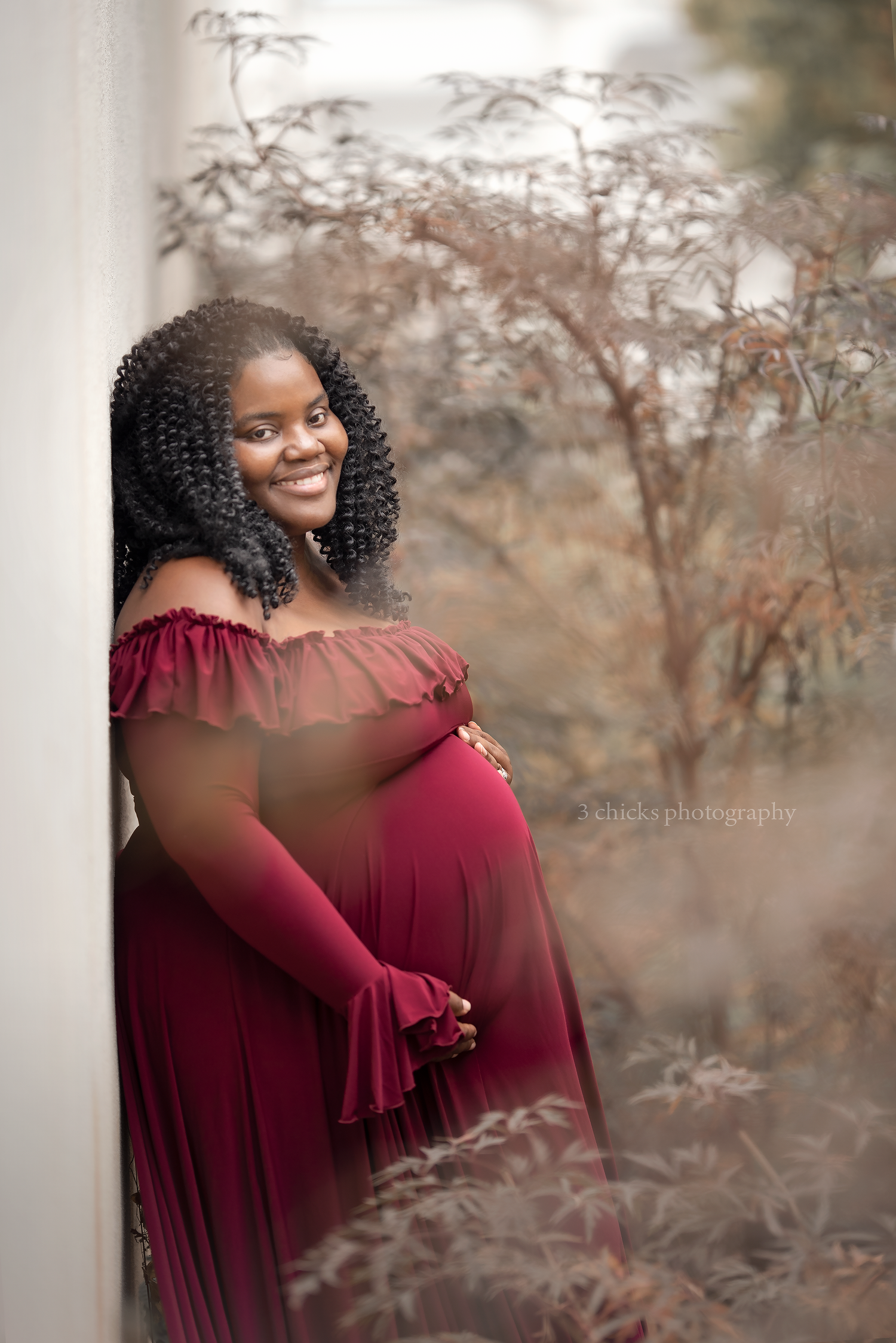 Smiling pregnant woman in burgundy off-shoulder maternity gown posing outdoors among autumn foliage.