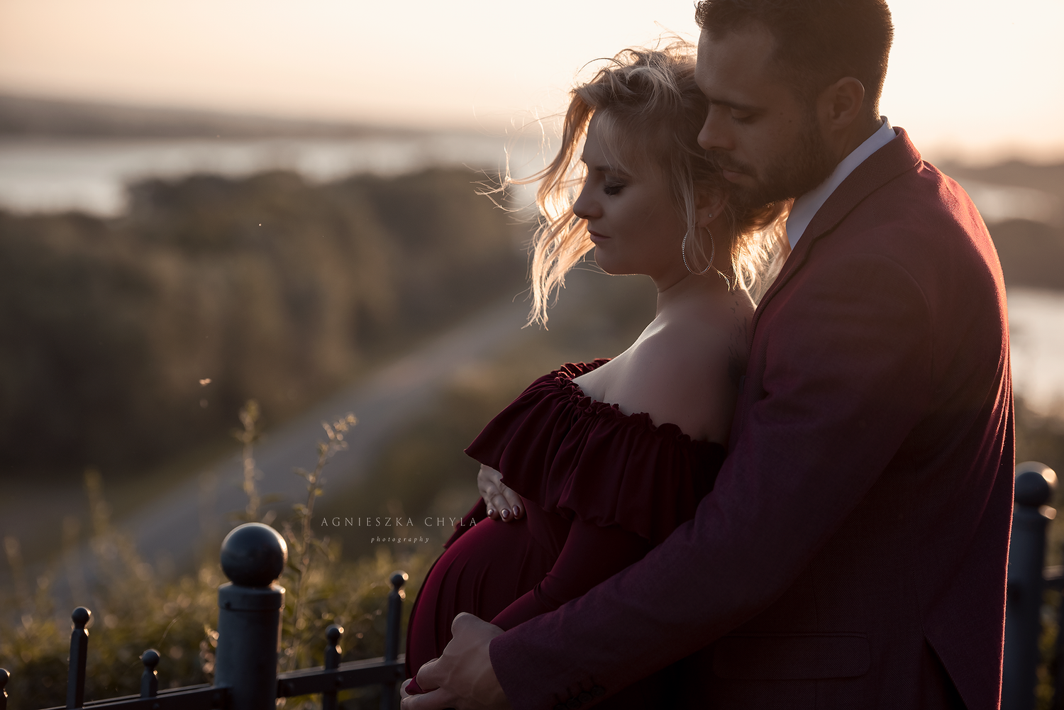 Couple embracing at golden hour sunset, woman in red off-shoulder gown, man in burgundy suit, romantic outdoor portrait.