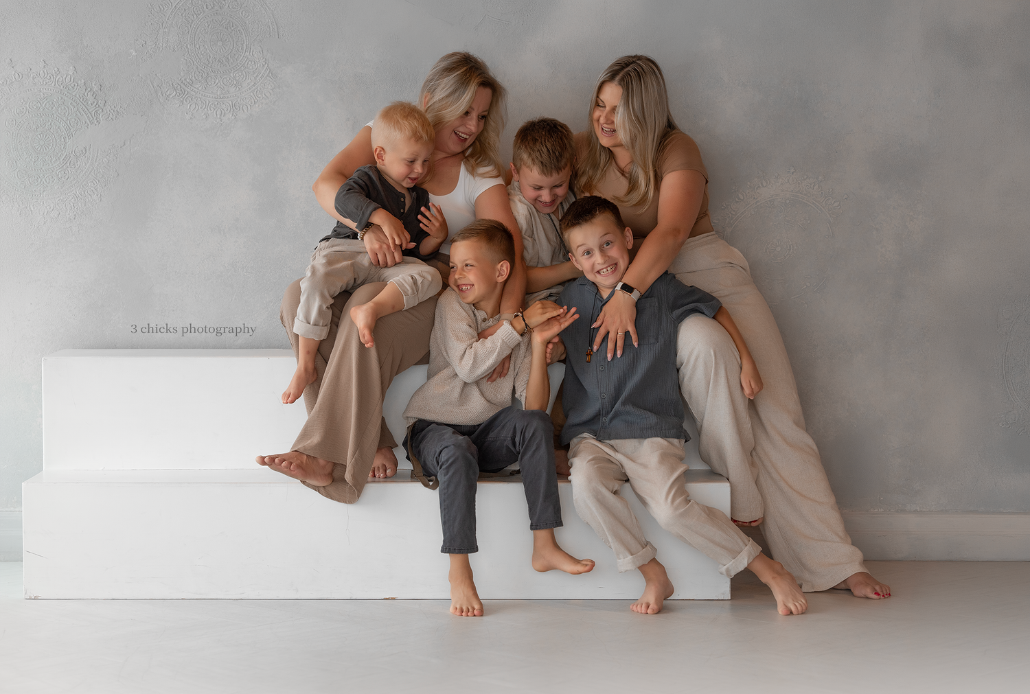 Family of five posing together on white steps against gray backdrop in coordinated neutral outfits for a professional photo.