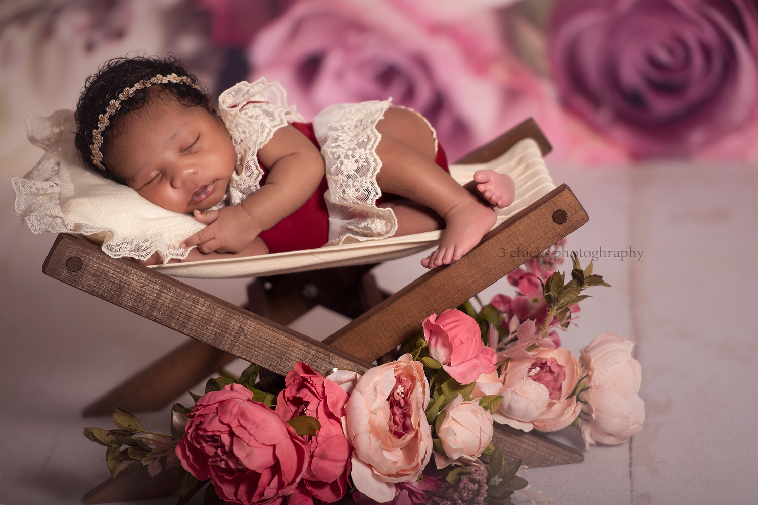 Newborn baby sleeping peacefully in a wooden chair surrounded by pink roses and peonies in a floral studio setting.