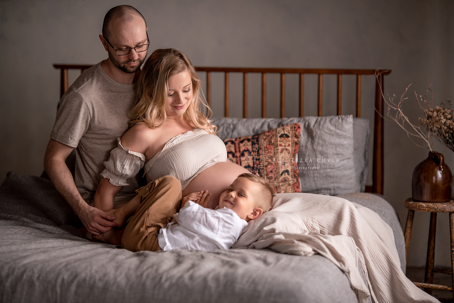 A couple sits on a bed, the pregnant woman cradling her belly while a young child rests against her bump.
