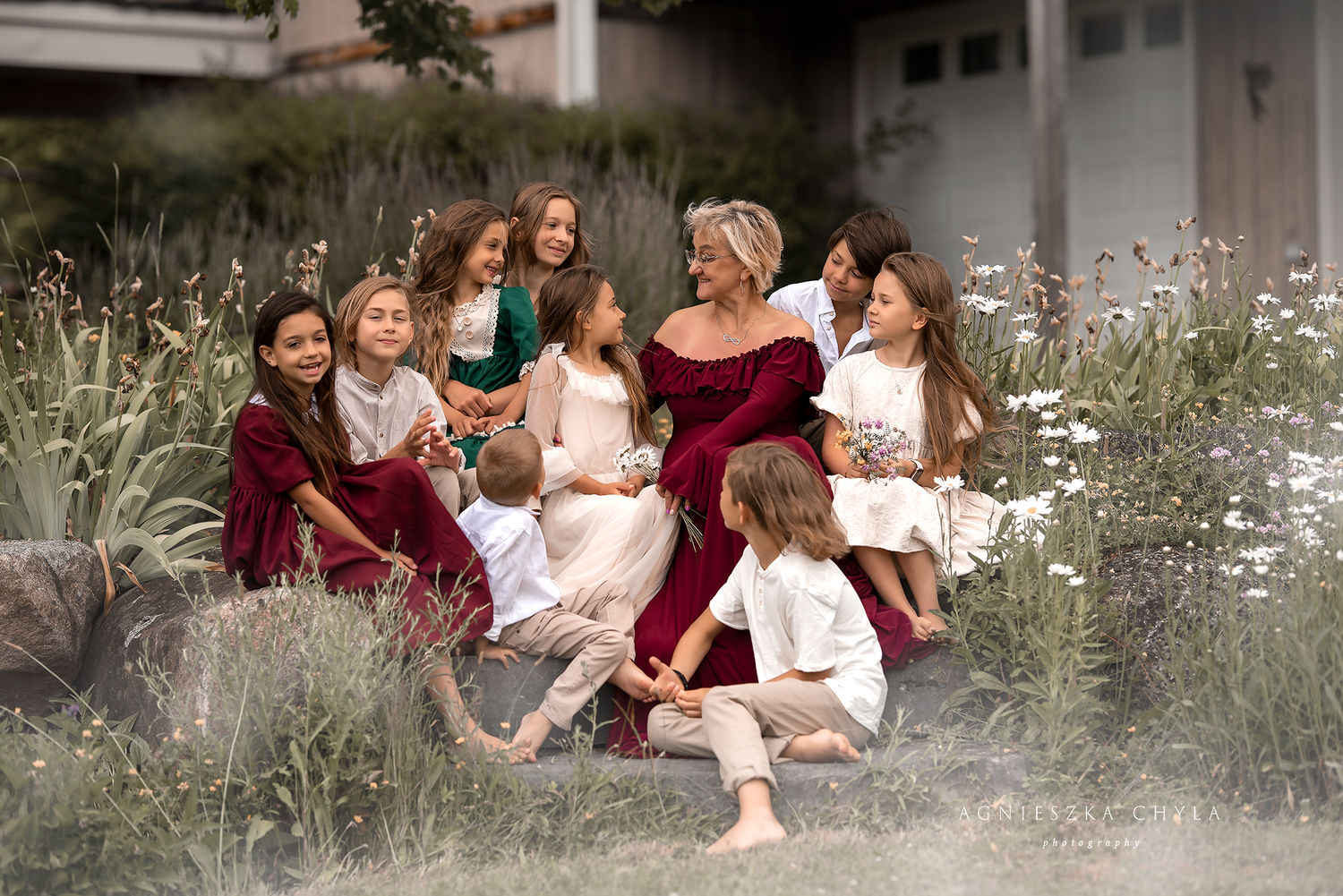 Blonde woman surrounded by group of children sitting on rocks in a garden setting.