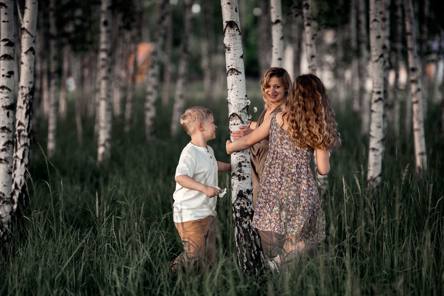 Curly-haired mother in floral dress playing with young son among white birch trees.