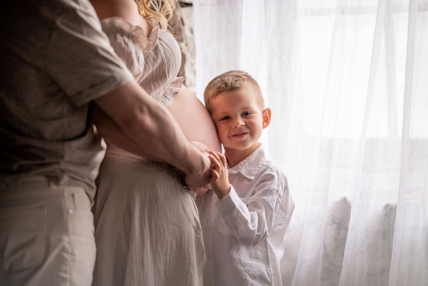 Smiling young boy in white shirt hugs pregnant mothers belly while standing by a bright window.