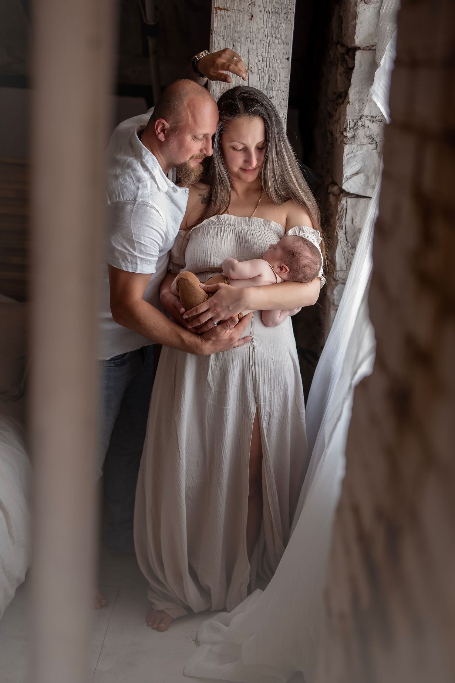A couple in white attire tenderly holds their newborn baby in a rustic indoor setting.