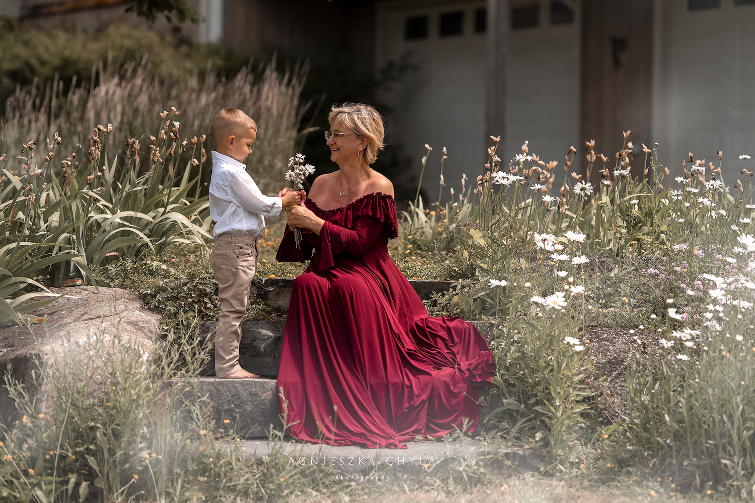 A woman in a red gown sits on steps with a young child offering flowers in a lush garden setting.