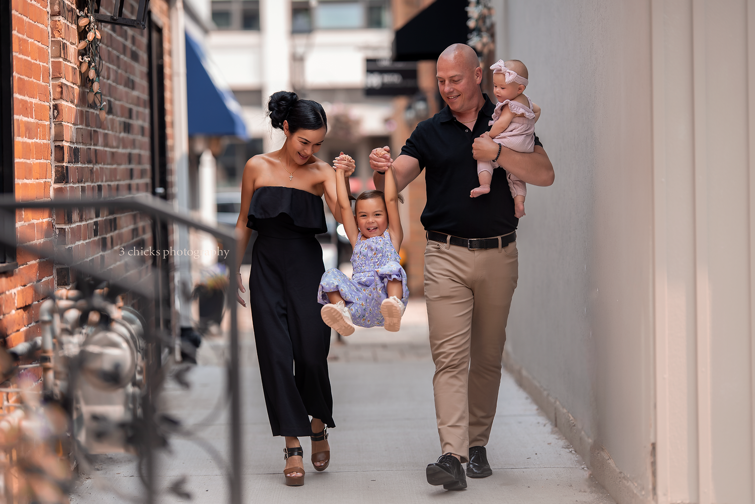 Happy family walking in urban alley, parents swinging toddler between them while dad holds baby.