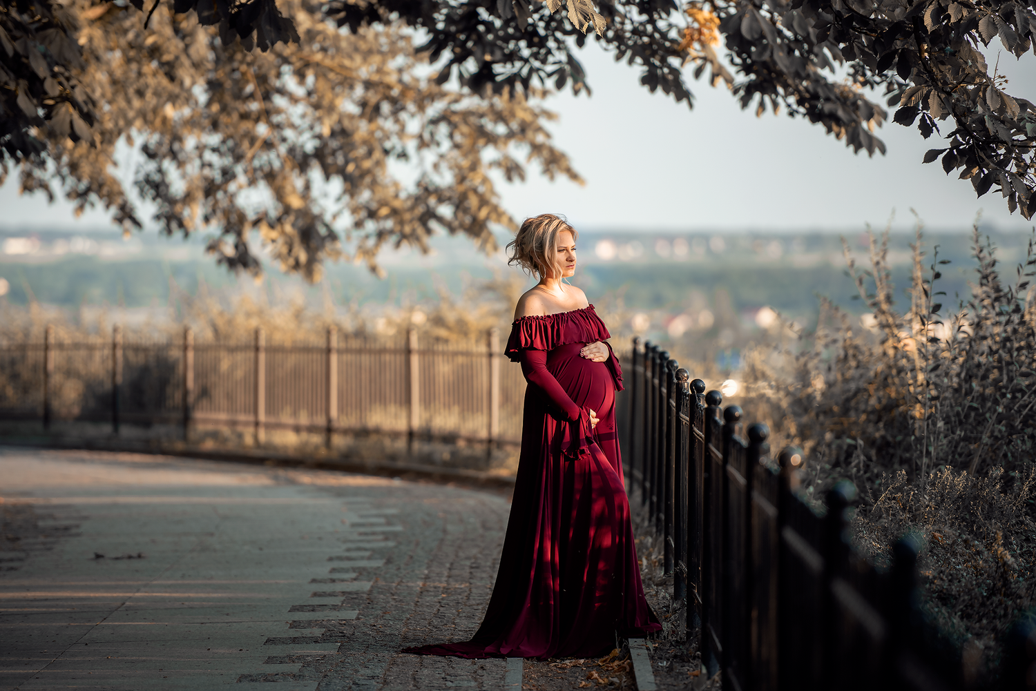 Pregnant woman in elegant burgundy off-shoulder gown poses gracefully along a scenic iron fence path surrounded by autumn trees.