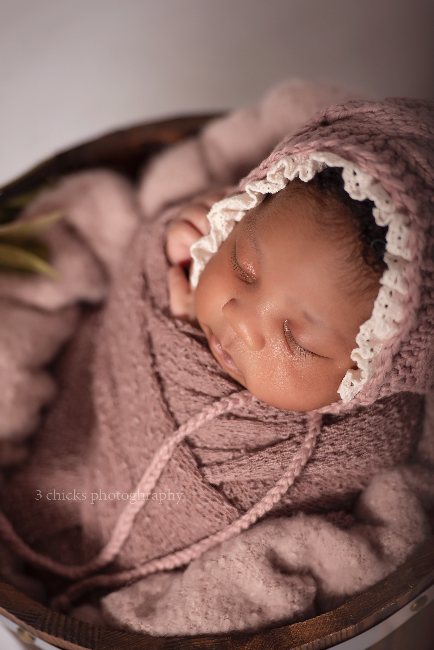 Newborn baby swaddled in dusty pink wrap and bonnet, sleeping peacefully in a wooden bowl by 3 Chicks Photography.