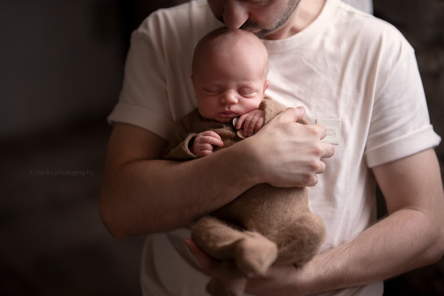 Father in white t-shirt tenderly kissing sleeping newborn baby wrapped in brown swaddle while holding a teddy bear.