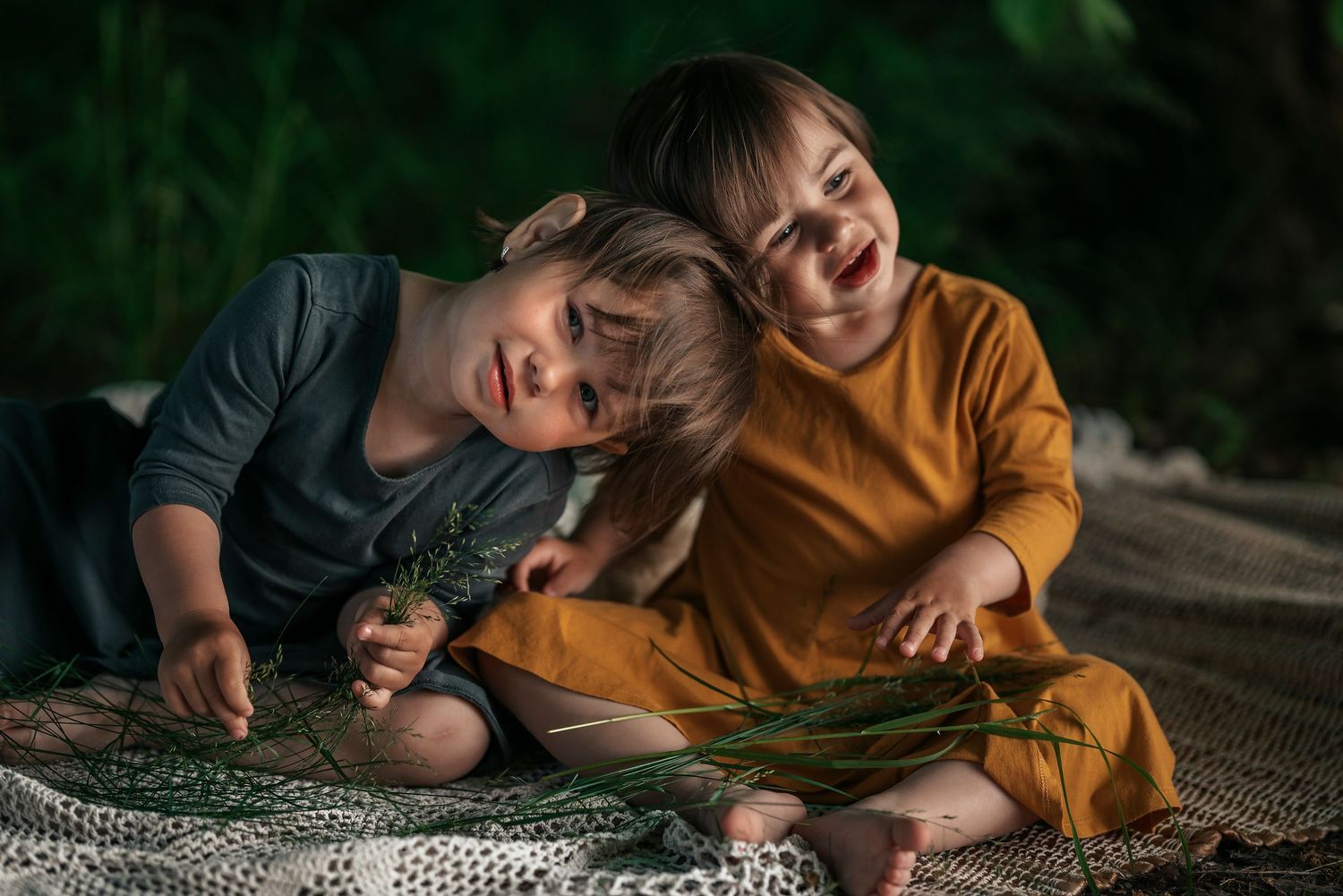 Two young children laughing and cuddling together on a blanket surrounded by green plants outdoors.