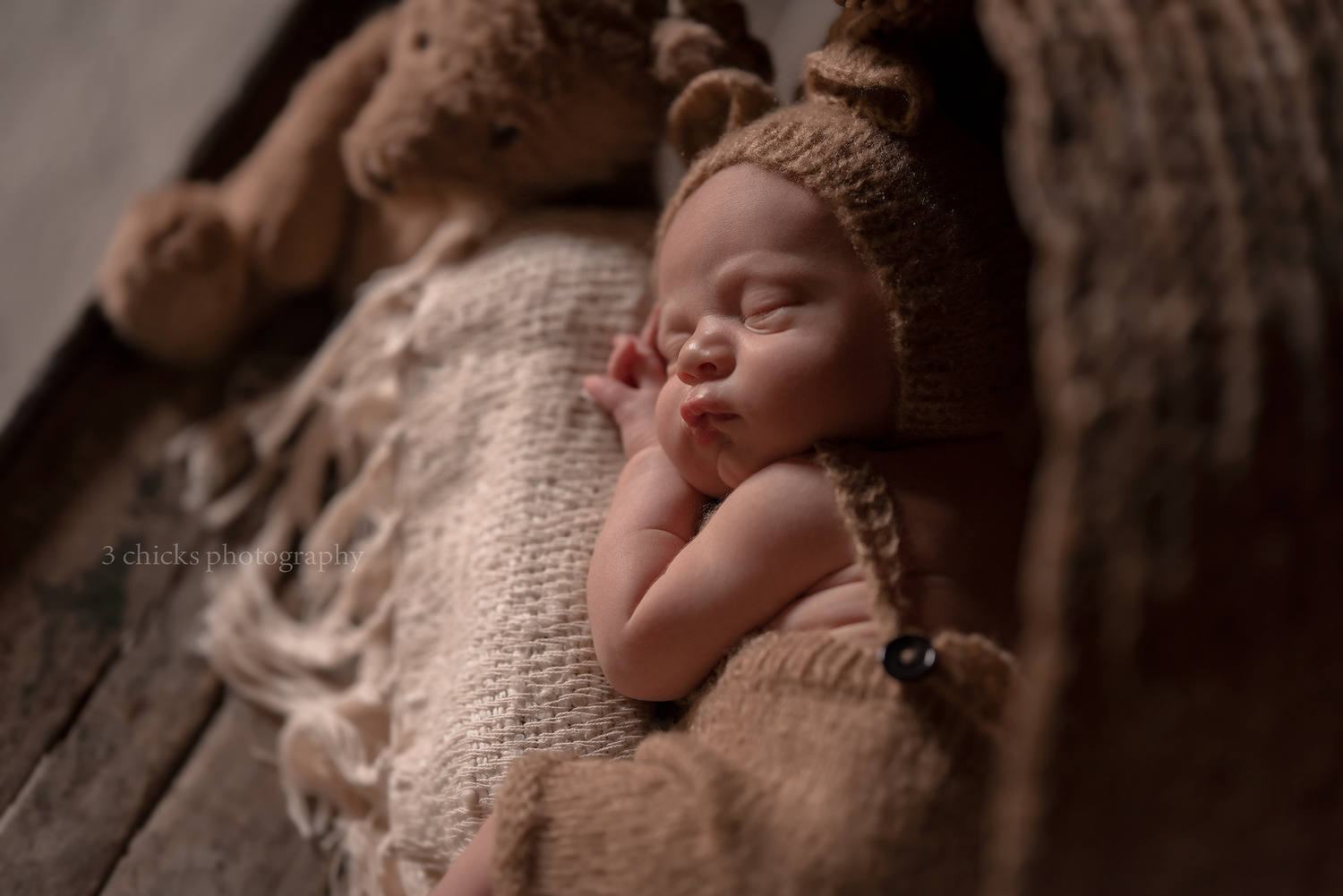 Sleeping newborn baby in brown knit bear hat nestled in rustic wooden bowl with soft blanket and teddy bear.