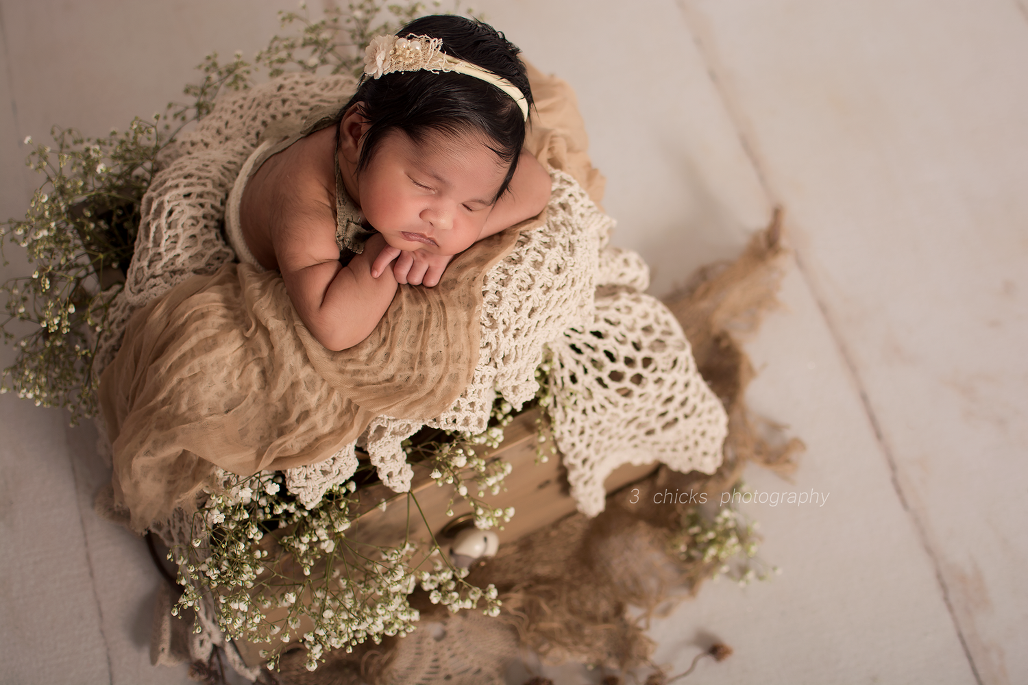 Newborn baby sleeping peacefully in a rustic bowl with lace wrap, floral headband, and delicate white flowers.