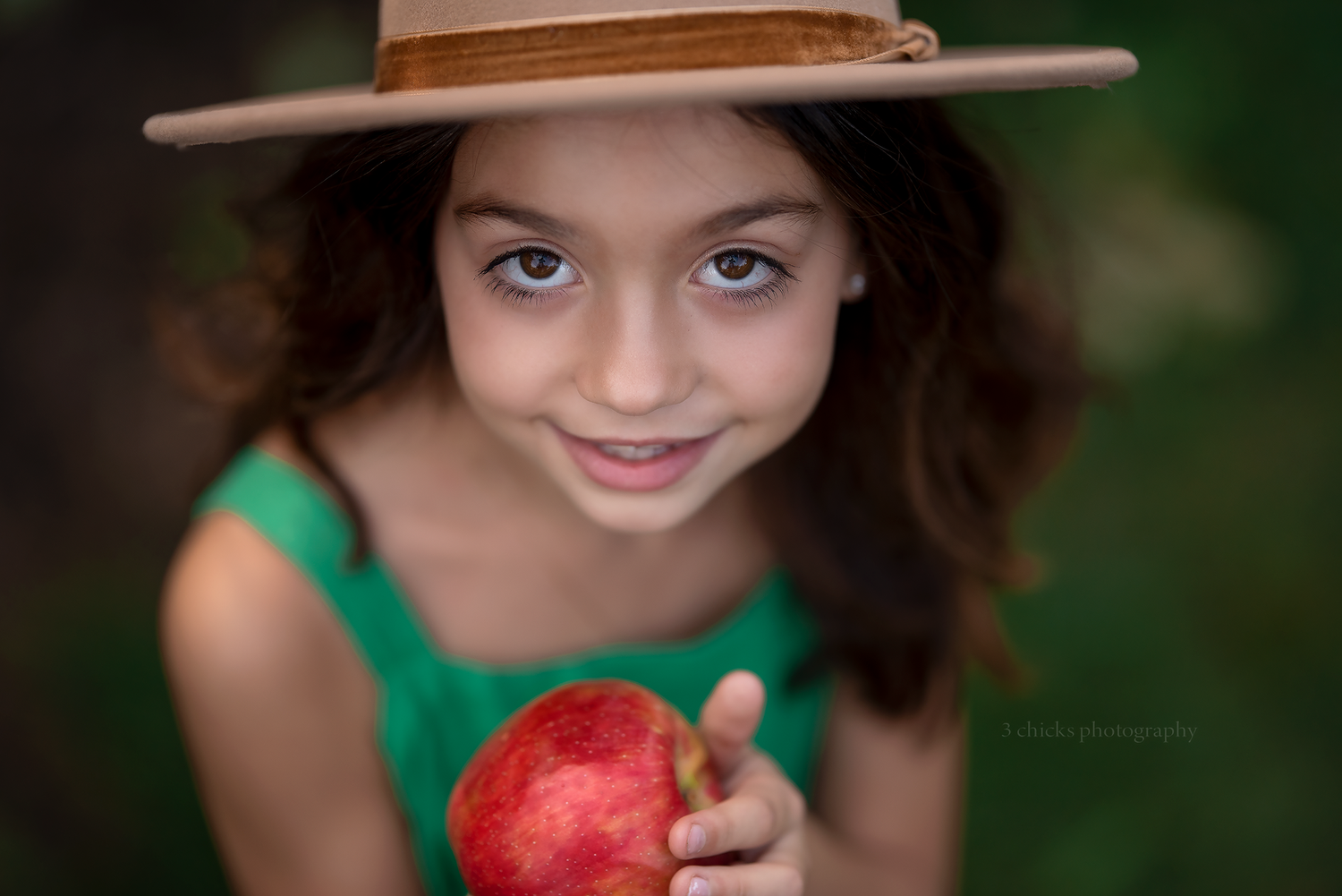 Cute girl with curly hair wearing a straw hat holding a red apple toward camera in garden.