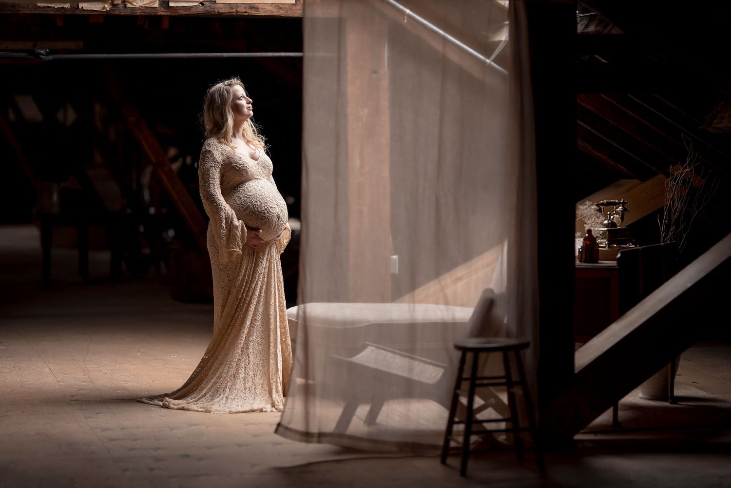 A pregnant woman in a lace gown stands in a dramatic beam of light in a dark studio.