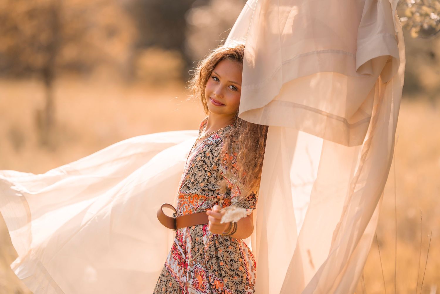 Young woman in floral dress holding flowers, standing in golden sunlit field with flowing white fabric.
