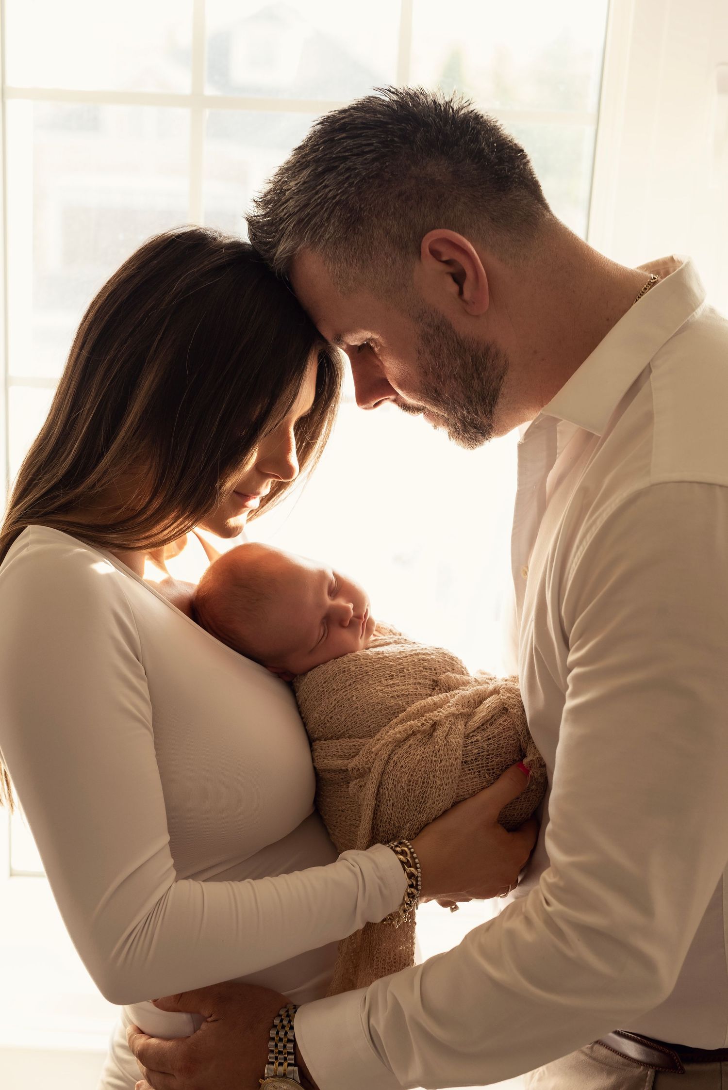 New parents tenderly hold their sleeping newborn baby wrapped in a knit blanket, foreheads touching in warm backlit light.