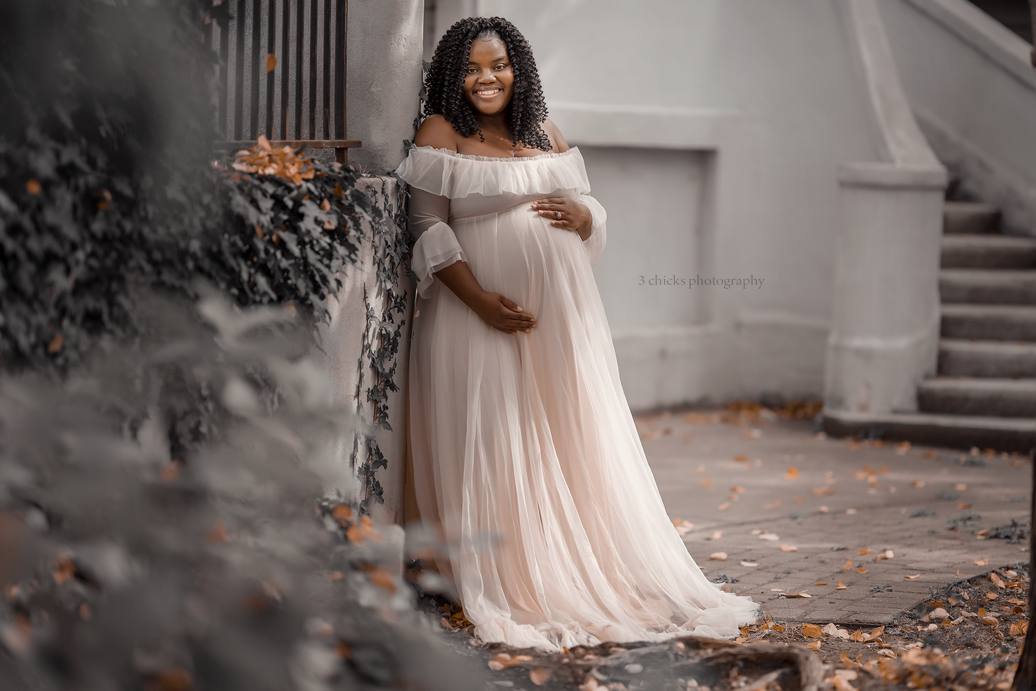 Smiling pregnant woman in elegant blush off-shoulder maternity gown posing outdoors against a white building.
