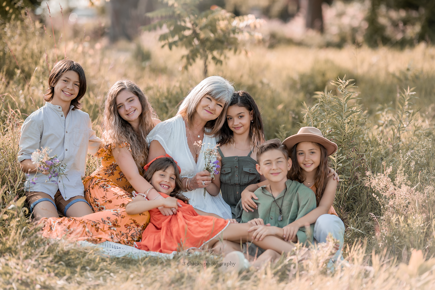 Group of six children and a woman with silver hair sitting together in a golden sunlit meadow.