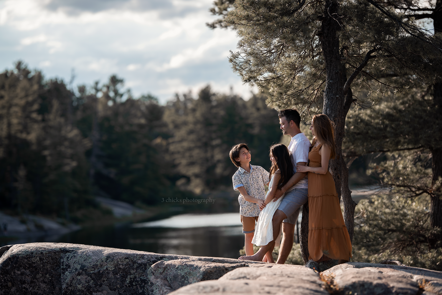 Family of four posing on rocky lakeside surrounded by pine forest in warm evening light.