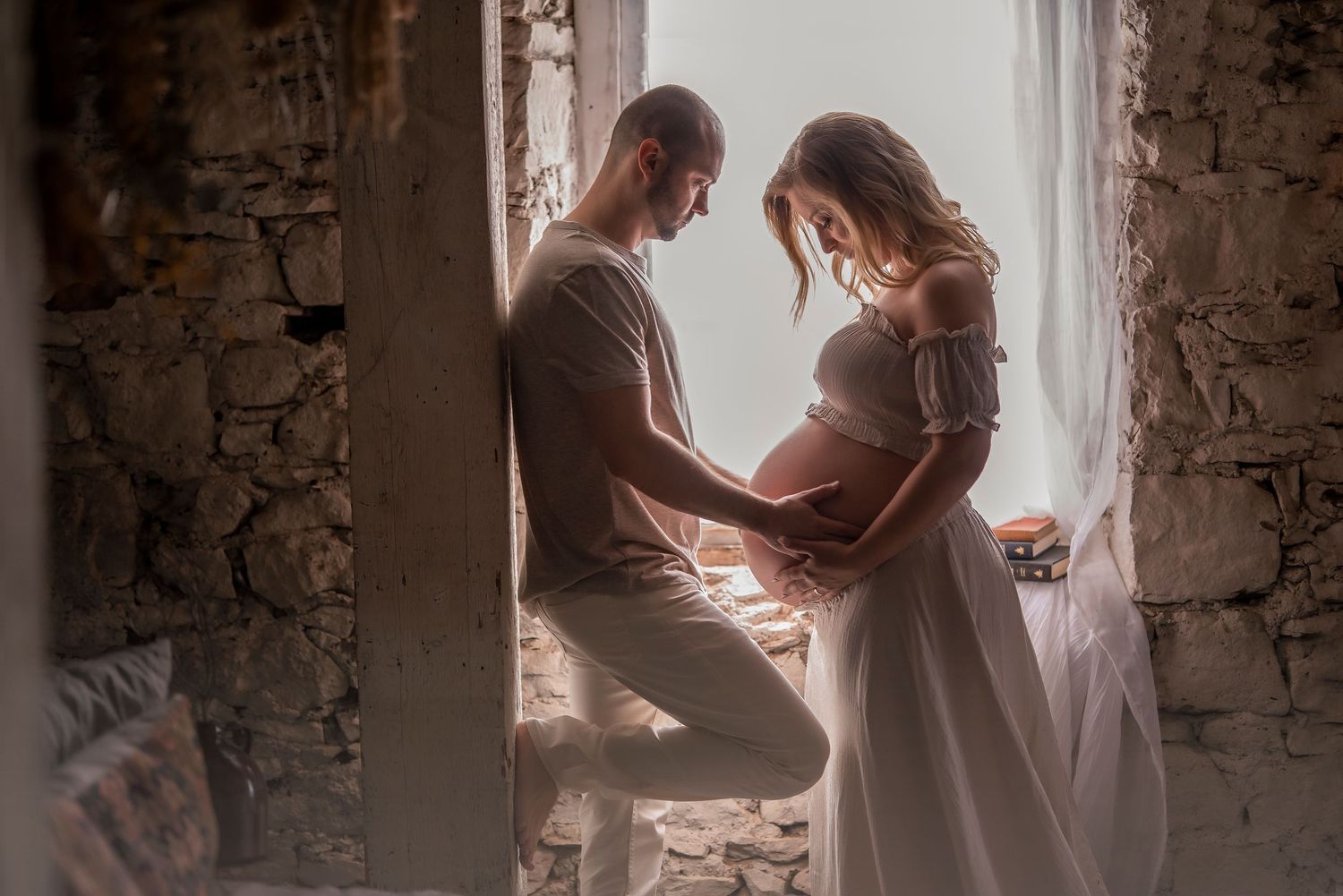 Couple tenderly holding pregnant belly together by rustic stone window in romantic maternity photo.