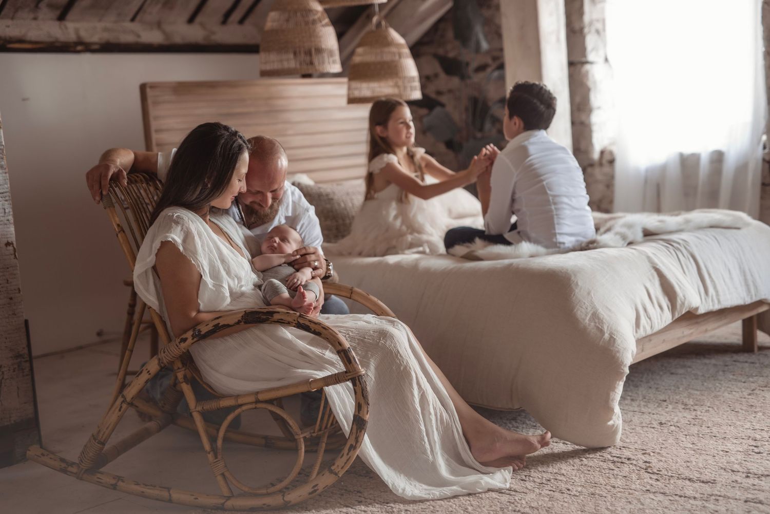 Mother in white dress sits in rattan rocking chair holding newborn while children play on bed behind her.