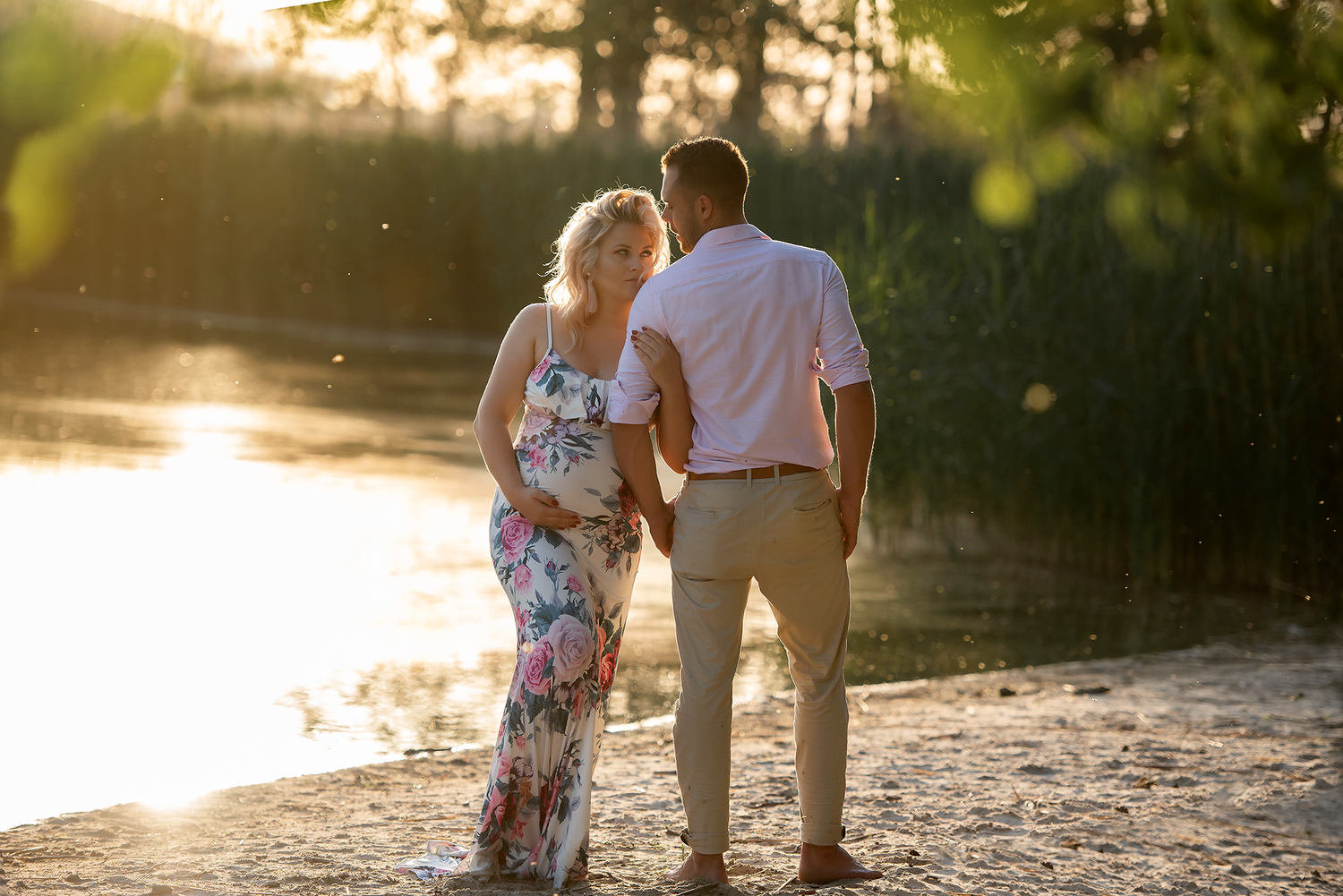 Couple holding hands by a sunlit lake shore at golden hour, woman in floral dress and man in white shirt.