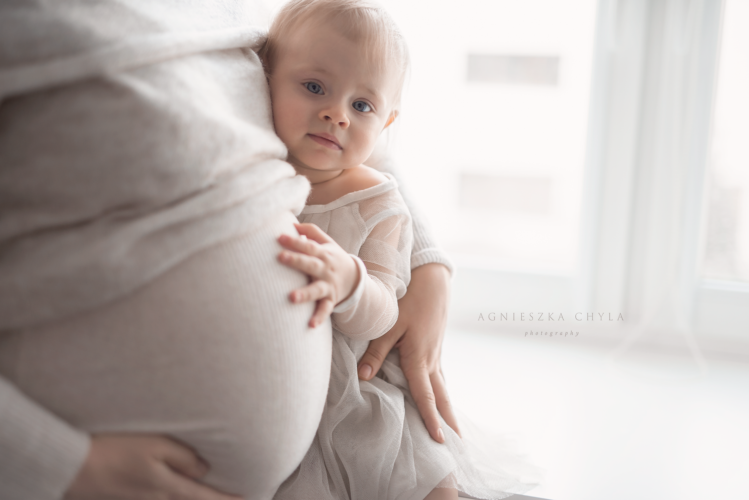 A curious baby in white onesie rests against a pregnant mother's belly in a bright, airy room.