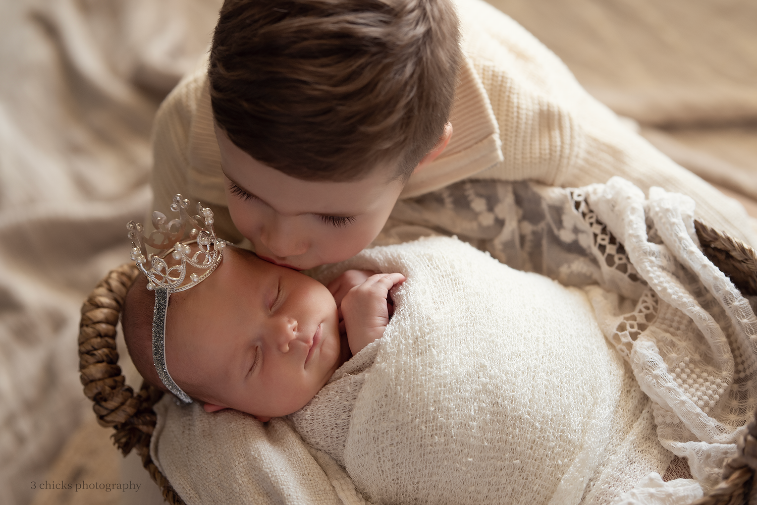 Older child gently kissing a swaddled newborn baby wearing a delicate crown headband in a cozy basket.