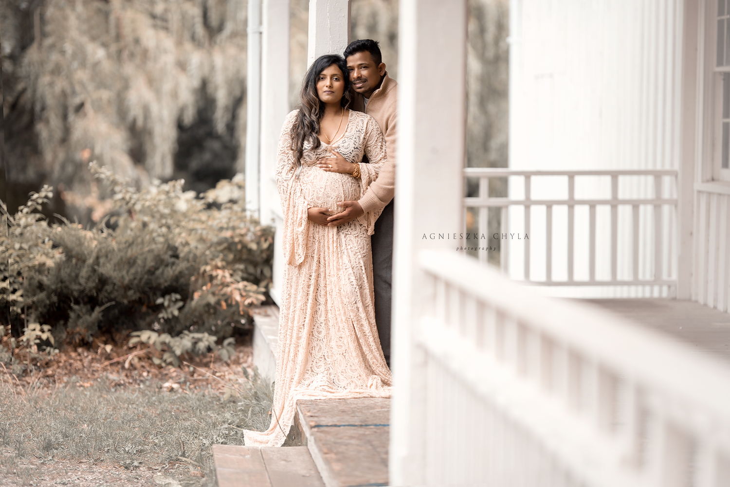 Couple in elegant attire poses on porch, woman in blush sequin maternity gown cradling baby bump.