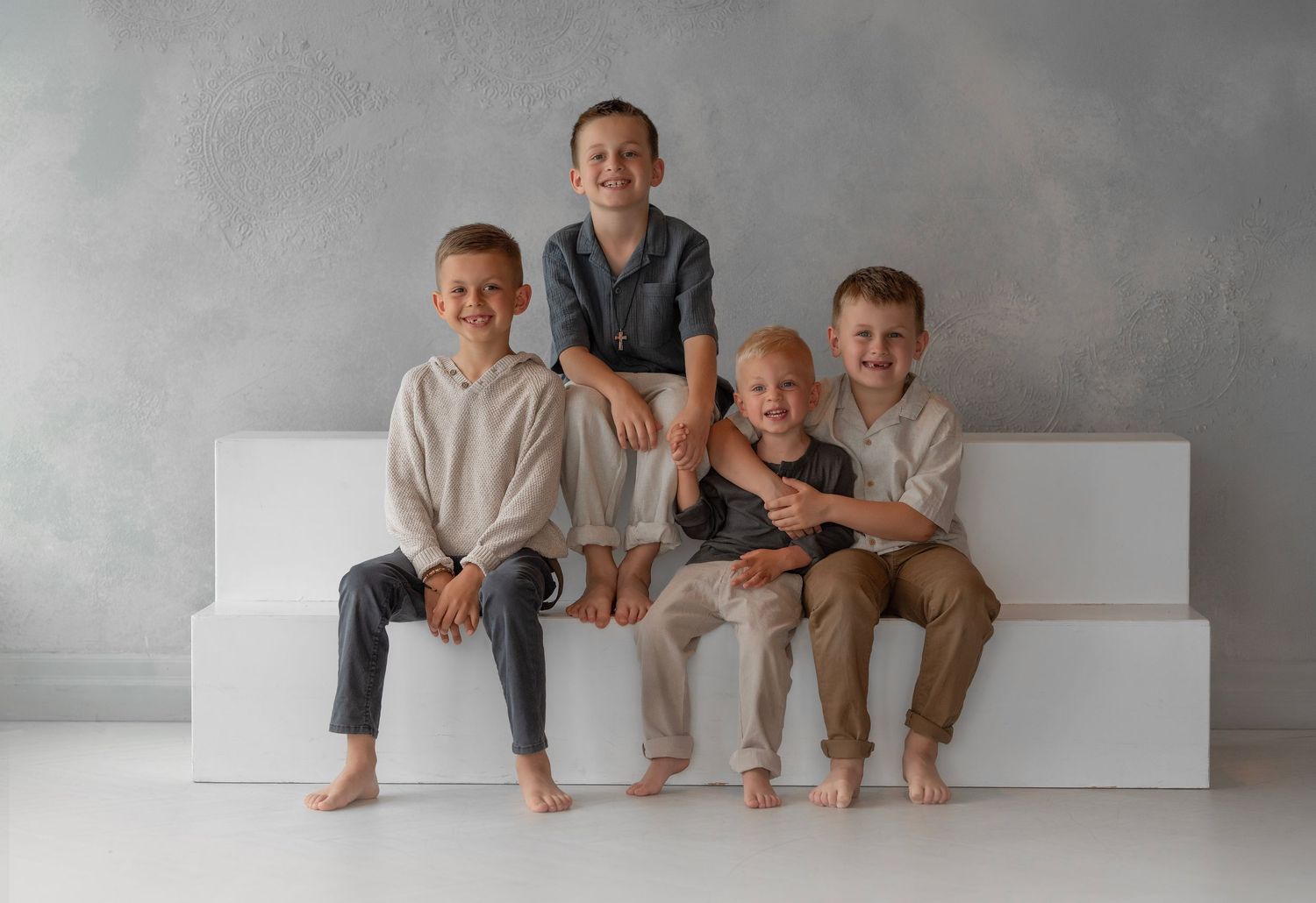 Four barefoot boys sitting and smiling on a white bench against a gray wall, dressed in neutral-toned casual outfits.
