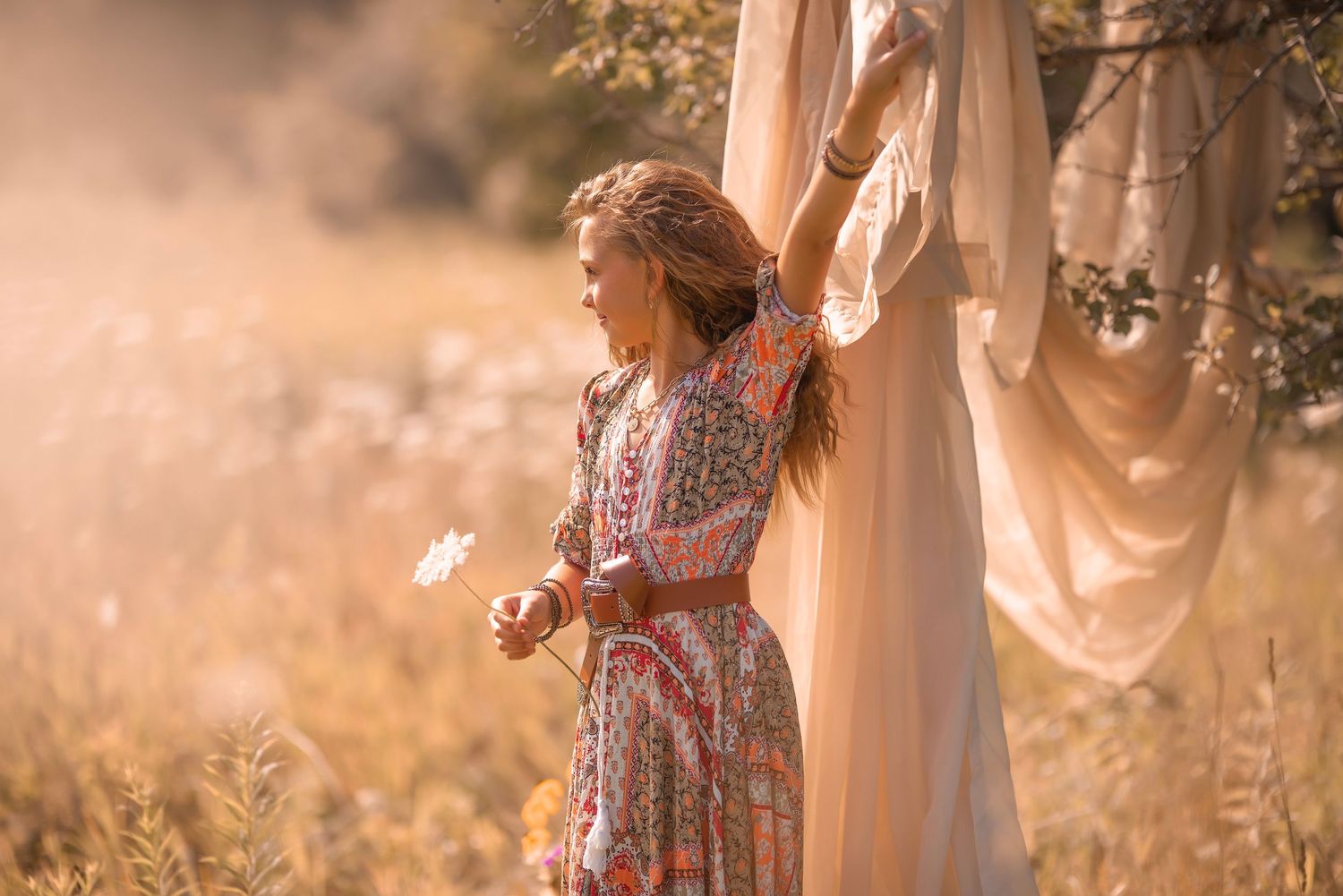 Woman in floral boho dress holding white flower in golden sunlit field near flowing fabric.