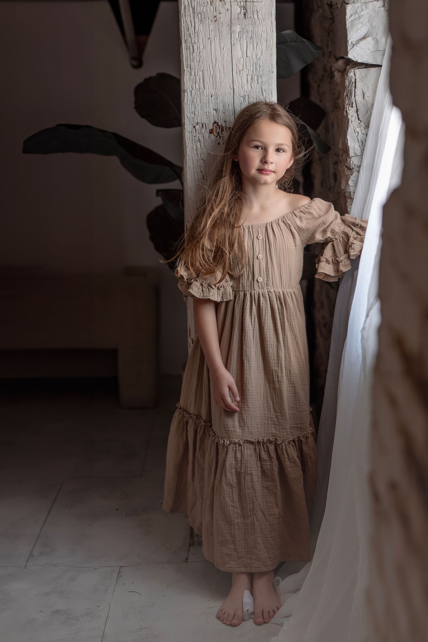 A young girl with long hair poses in a tan boho dress in a rustic indoor setting.