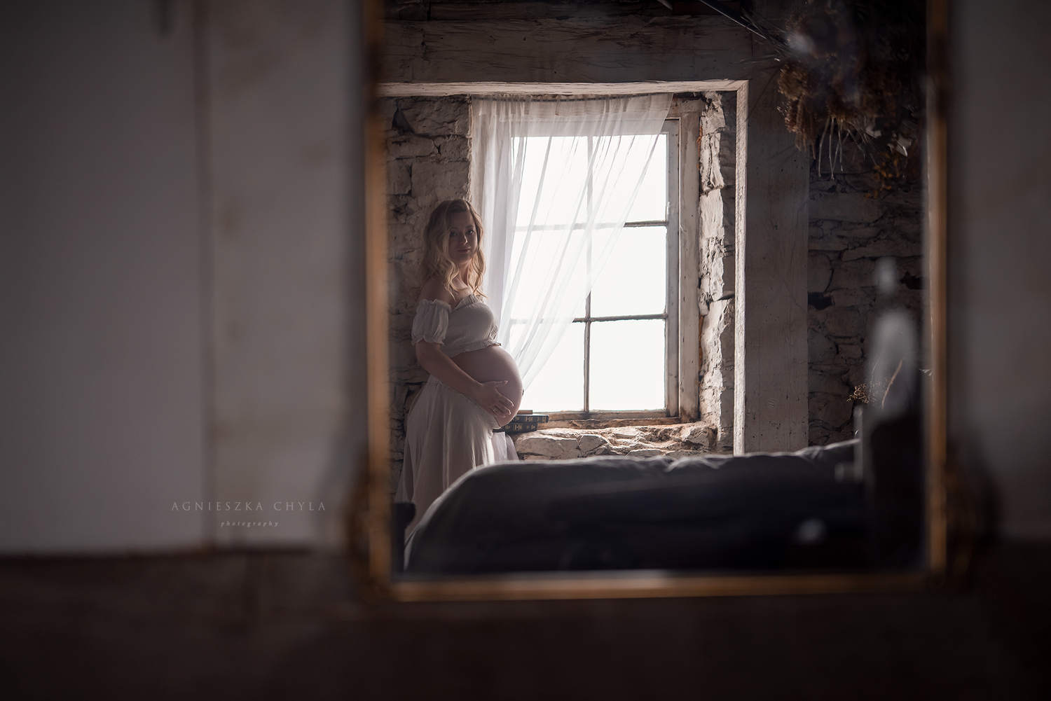 Pregnant woman in white dress standing by a rustic window, reflected in a gold-framed mirror in a vintage room.