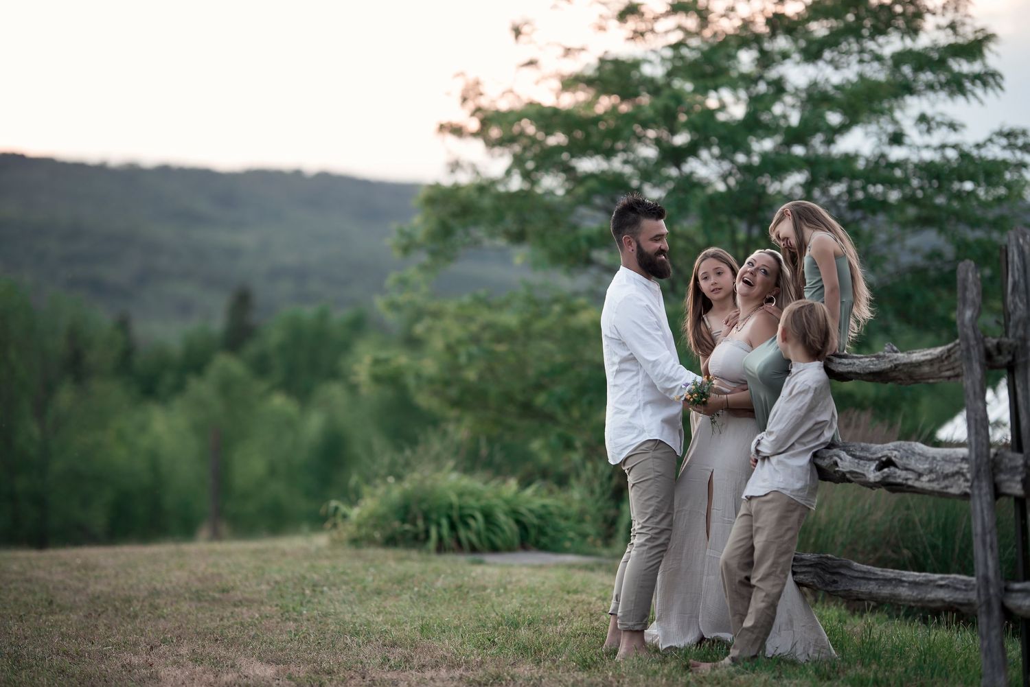 Happy family of four posing near a wooden fence in a lush green countryside setting.