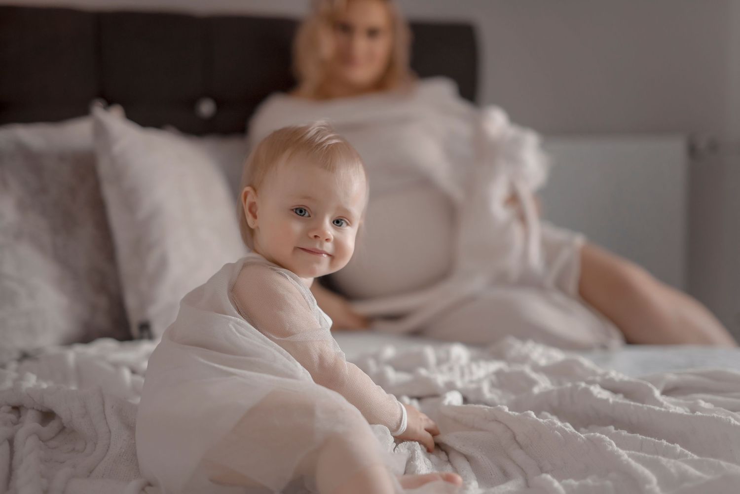 Baby in white onesie sitting on bed smiling, with pregnant blonde woman resting in soft white bedroom background.
