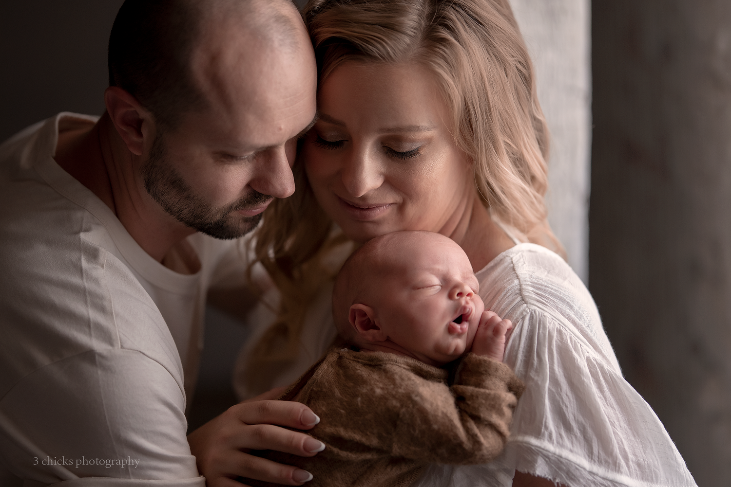 New parents tenderly holding their sleeping newborn baby in a soft, intimate studio portrait photo.