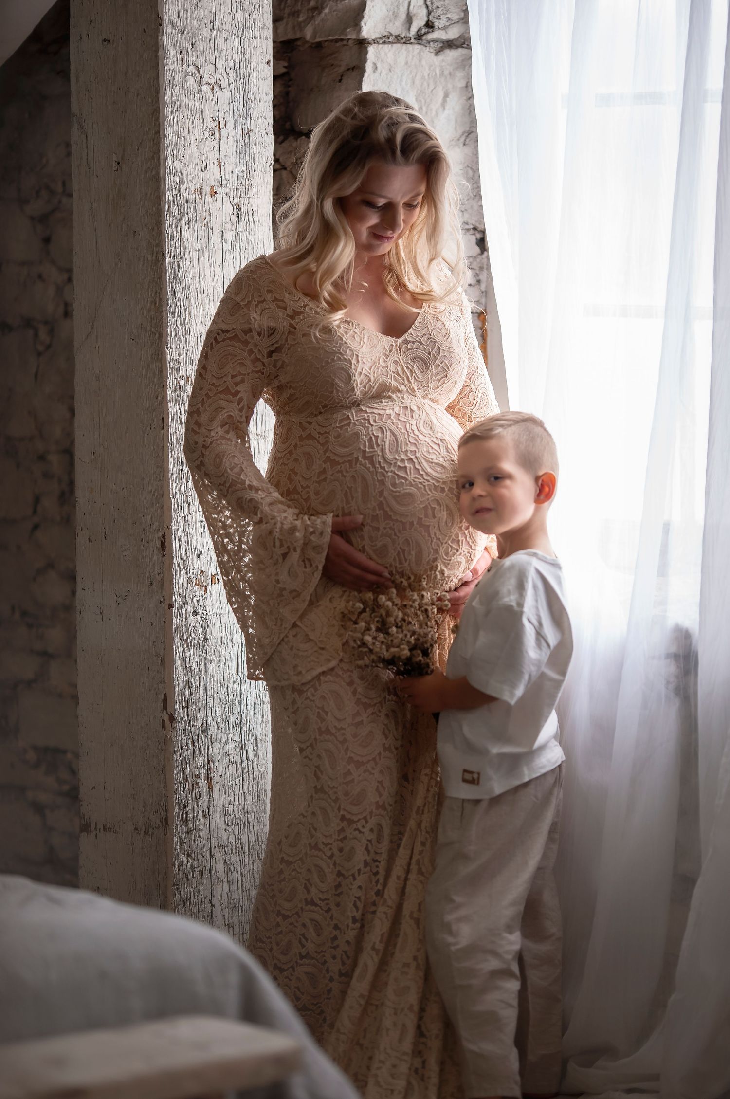 A pregnant woman in a lace gown stands by a window as a young boy hugs her baby bump.