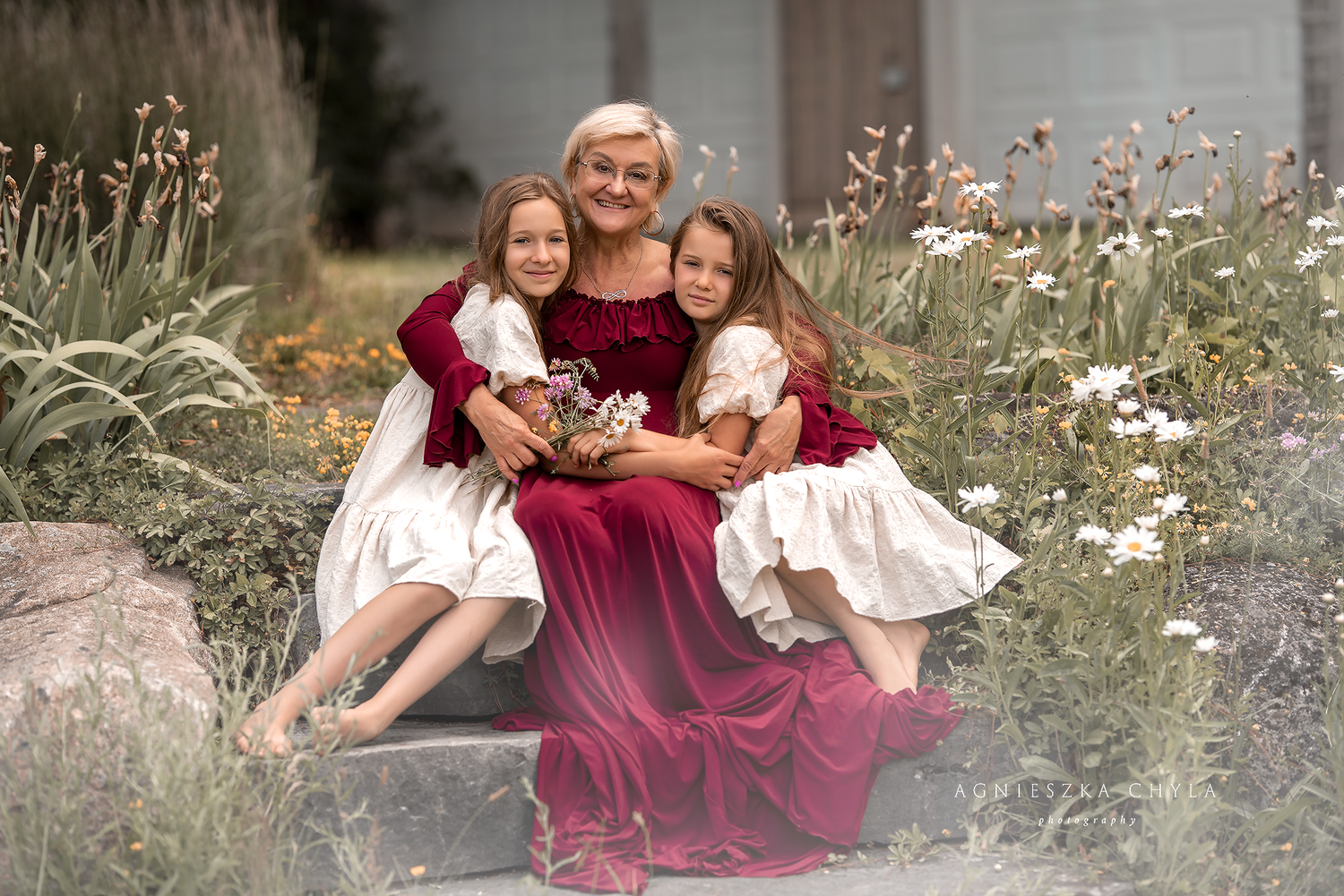 Grandmother in red dress sitting with two young granddaughters in flower garden.