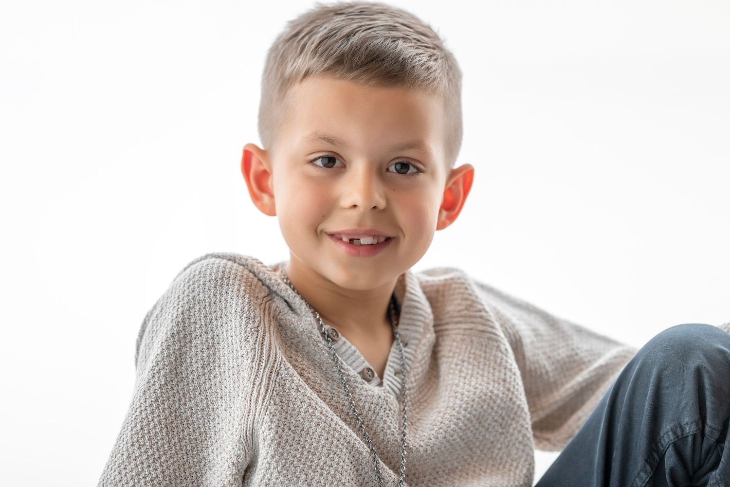 Smiling young boy with short blonde hair wearing a gray knit cardigan, posed against a white background.