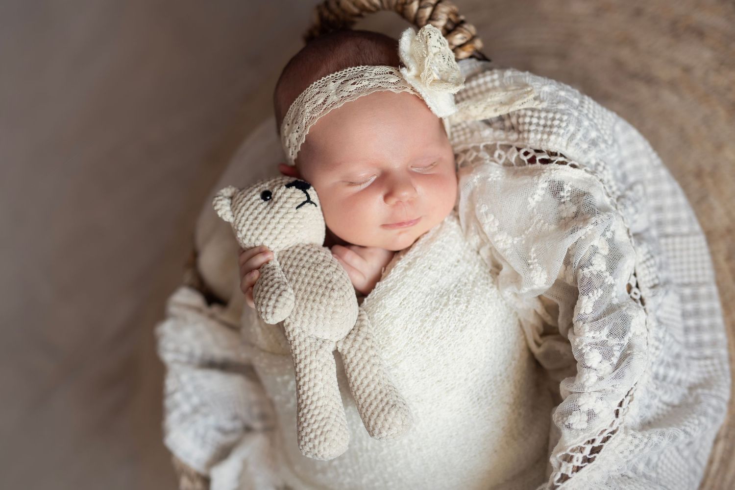Sleeping newborn baby wrapped in white lace holding a knit teddy bear with a floral headband in a basket.