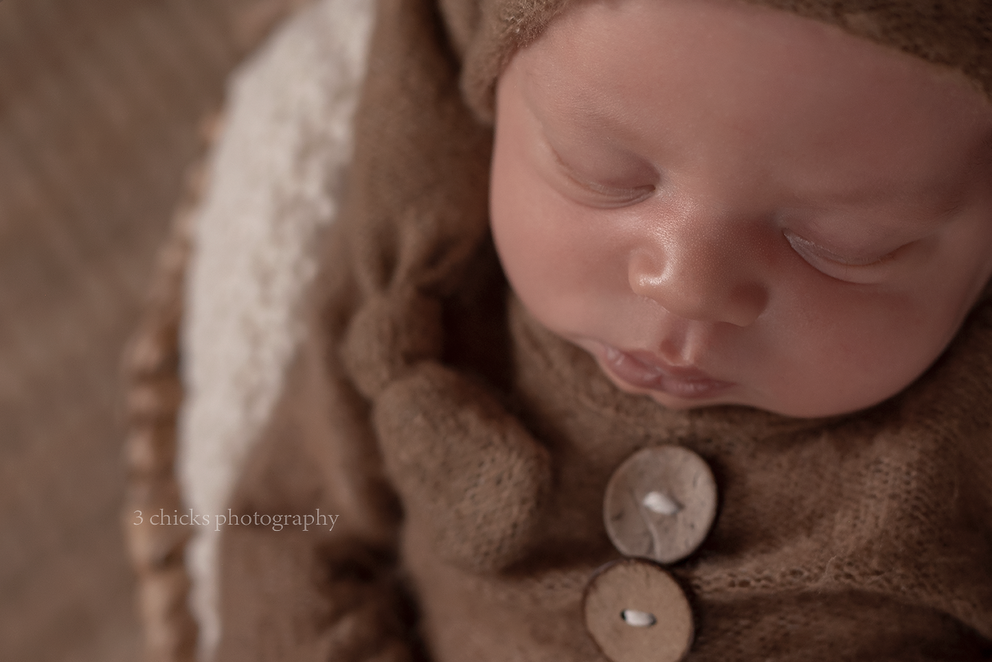 Sleeping newborn baby wrapped in soft brown knit blanket with wooden buttons, wearing a cozy brown hat.
