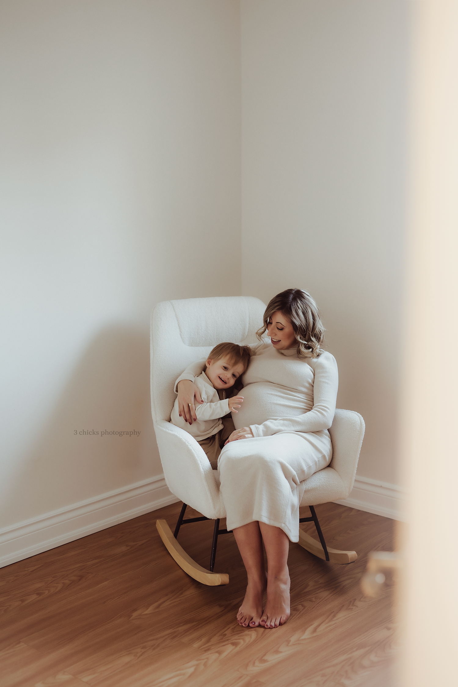 Pregnant woman in white dress sitting in rocking chair with toddler, maternity photography session in bright minimalist room.