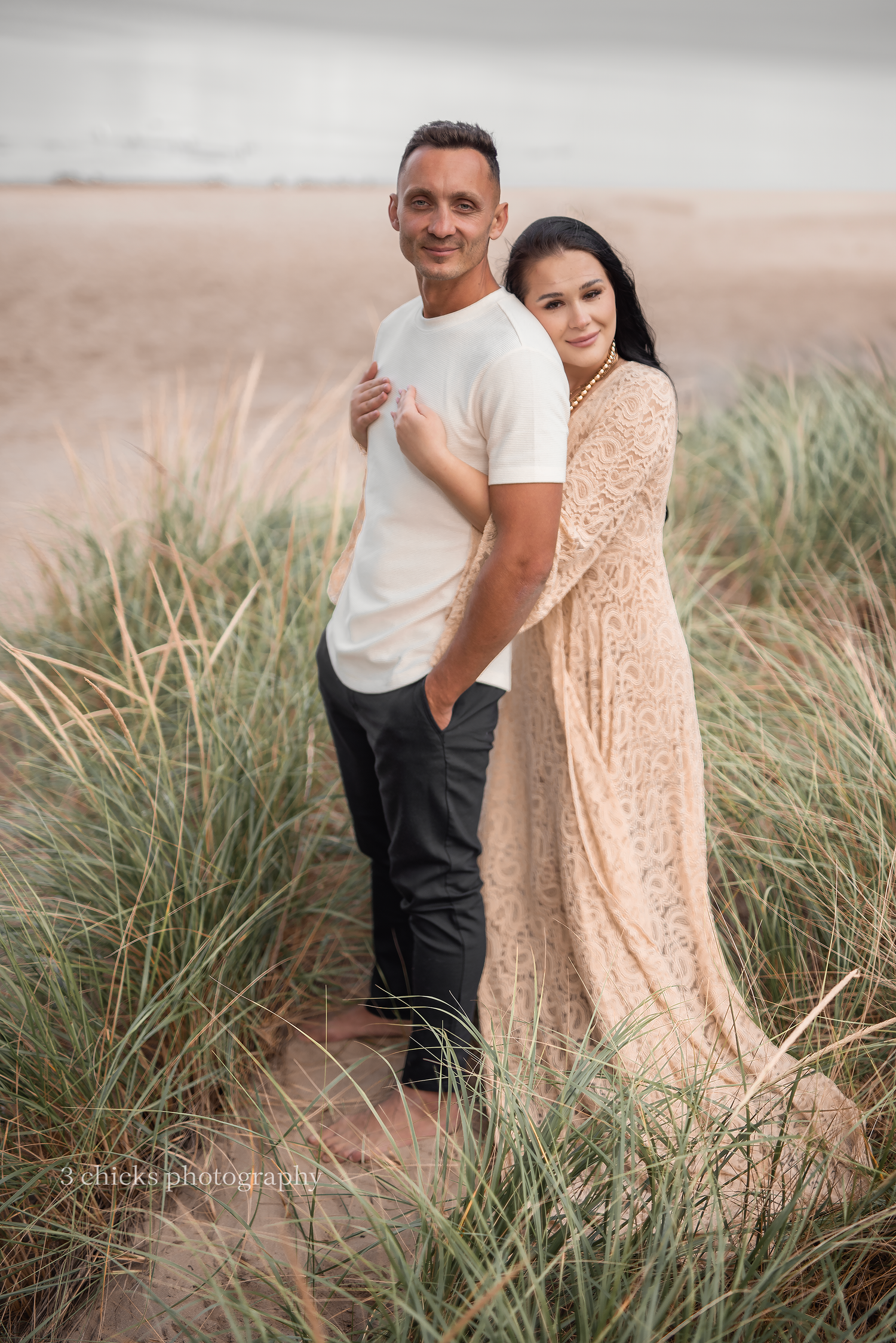 Couple posing together on a beach , woman in lace dress, man in white tee and black pants. Couple photography session Toronto
