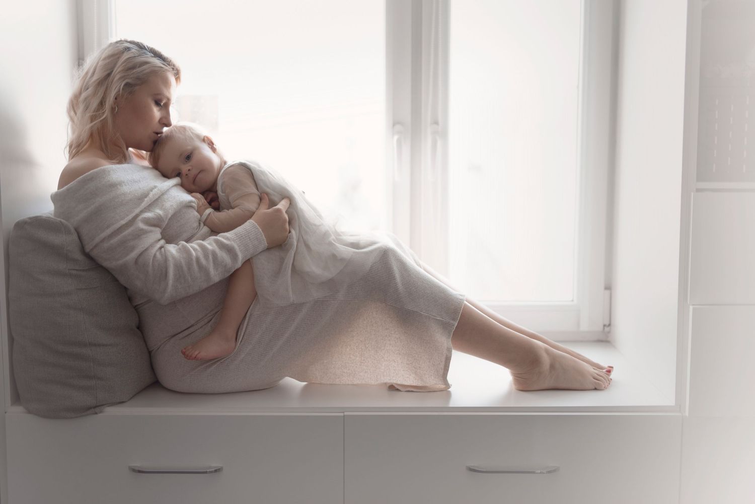 mother in a white robe sits on a window seat cradling her newborn baby in soft natural light. Maternity and newborn photography Cambridge